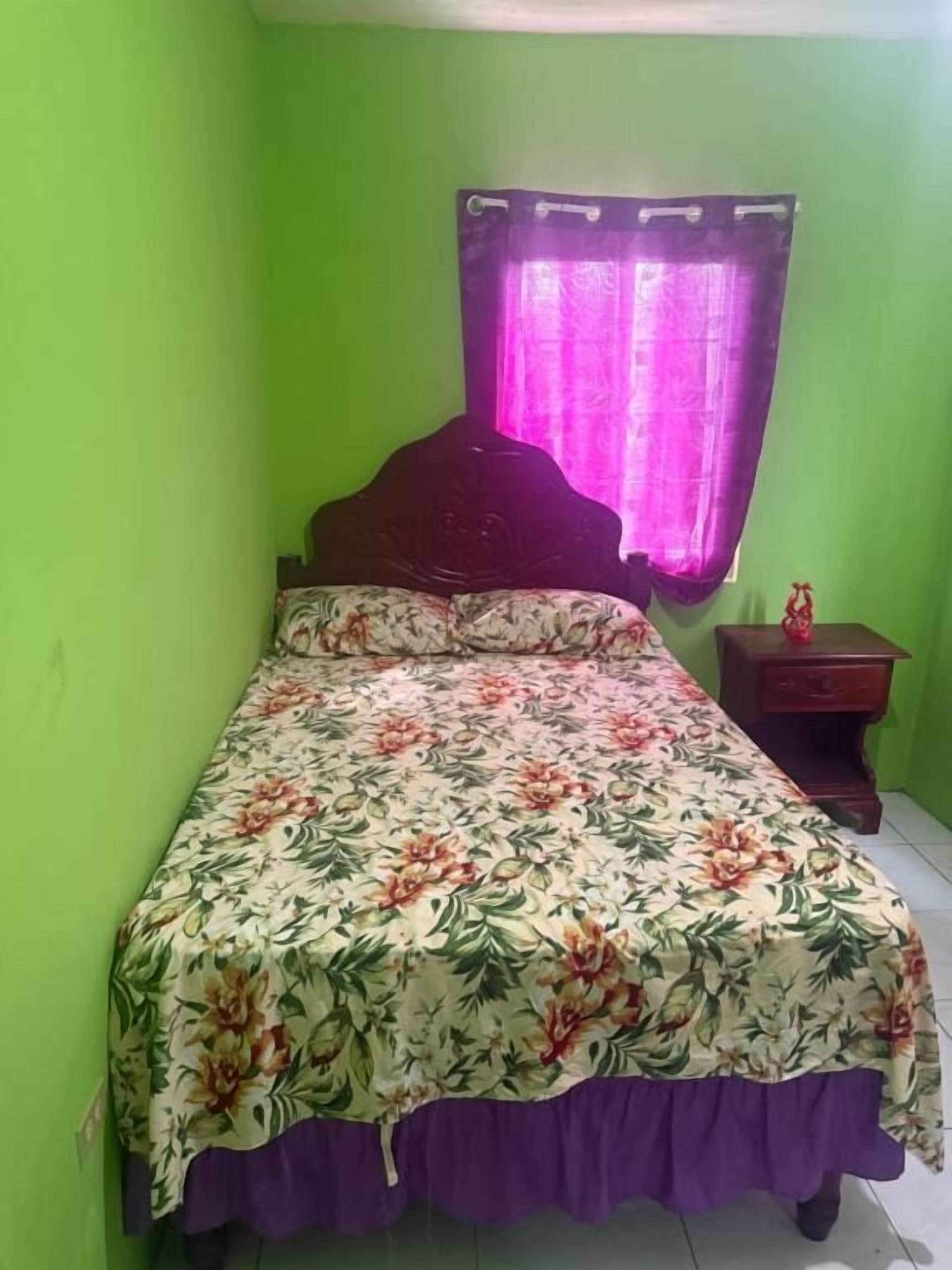 Una cama individual con colcha floral y detalles morados, una mesita de noche de madera con una pequeña estatua, y una ventana con cortina morada.