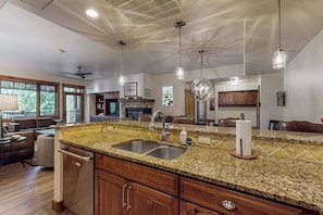 A kitchen with granite countertops, stainless steel sinks, wooden cabinets, pendant lighting, and a view of the living area. - Stylish condo near slopes with fireplace, balcony, grill, pools, & hot tubs (Steamboat Springs)