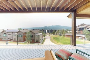 Ein überdachter Außenbereich mit Holzterrasse, einer Couch und Blick auf Gebäude und Berge.