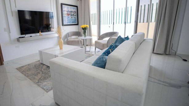 A modern living room with a white sofa, a flat-screen TV mounted on the wall, a small round table, and a vase with yellow flowers. - Perfect 3-bedroom Modern and Spacious Beach House (Manabí)