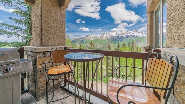 A balcony with a grill, two chairs, and a table overlooking a mountainous landscape.