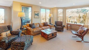 A spacious living room with a leather sofa, patterned armchairs, a wooden coffee table, and a view of mountains through large windows.