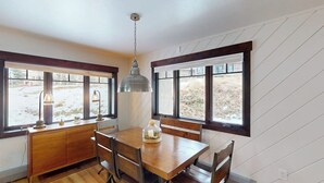 Un comedor con una mesa y sillas de madera, una lámpara colgante, un armario de madera y ventanas amplias con vistas al exterior.