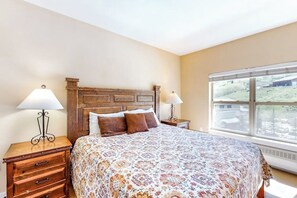 A bedroom with a wooden headboard, two bedside tables with lamps, a large bed with a patterned bedspread, and a window with a view of a landscape. - Base Camp #41 ski-in/ski-out condo with valley and pool views, ski locker (Granby)