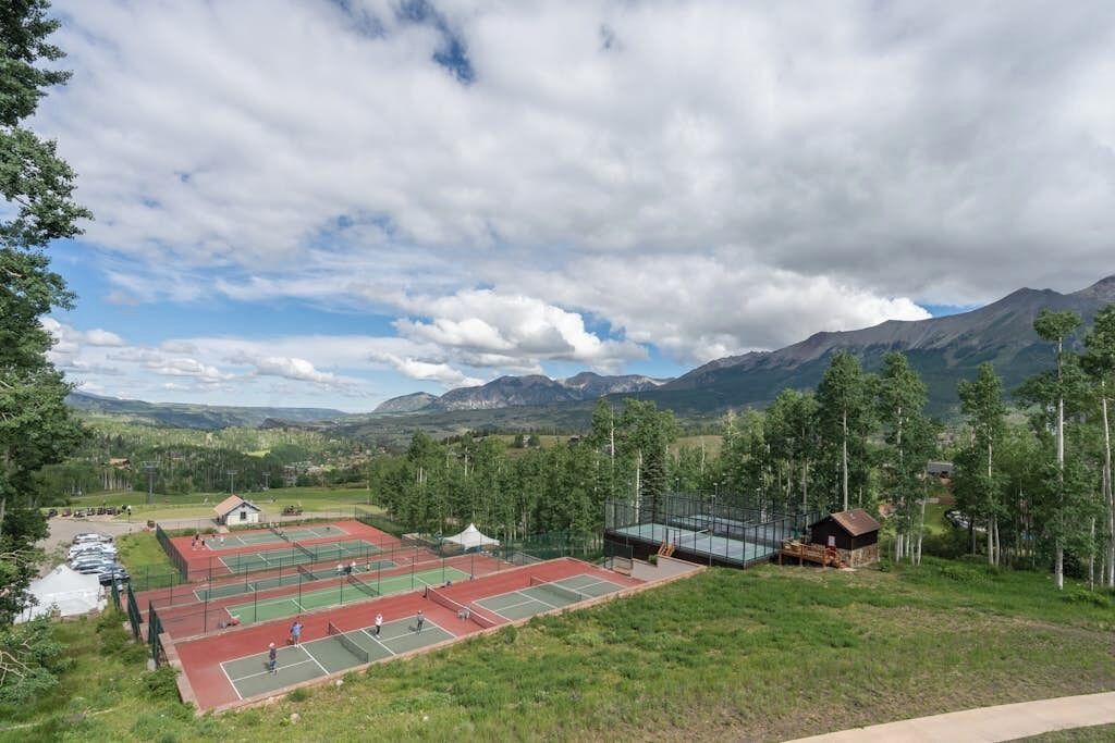 A tennis court complex with multiple courts, a small building, and a mountain backdrop.
