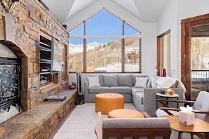 Ein gemütliches Wohnzimmer mit Kamin aus Stein, einem grauen Ecksofa und einem großen Fenster mit Blick auf die Berge.