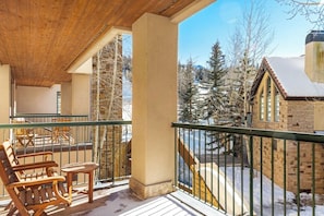 Eine Holzdar Terrasse mit einer Bank und einem Tisch, ein Steinhaus mit Schornstein und Blick auf schneebedeckte Bäume.