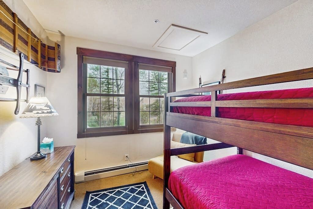 A bunk bed with a red mattress, a wooden dresser, a lamp, and a window with a view of trees.