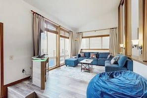 Ein Wohnzimmer mit einer Couch, einem Hocker und einem abgedeckten Sitzsack. Es steht ein Schreibtisch mit Computer und Lampe darin.