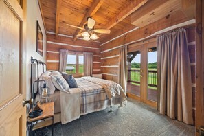 A log cabin bedroom with a bed, a ceiling fan, a nightstand with a lamp, and a view of the outdoors.