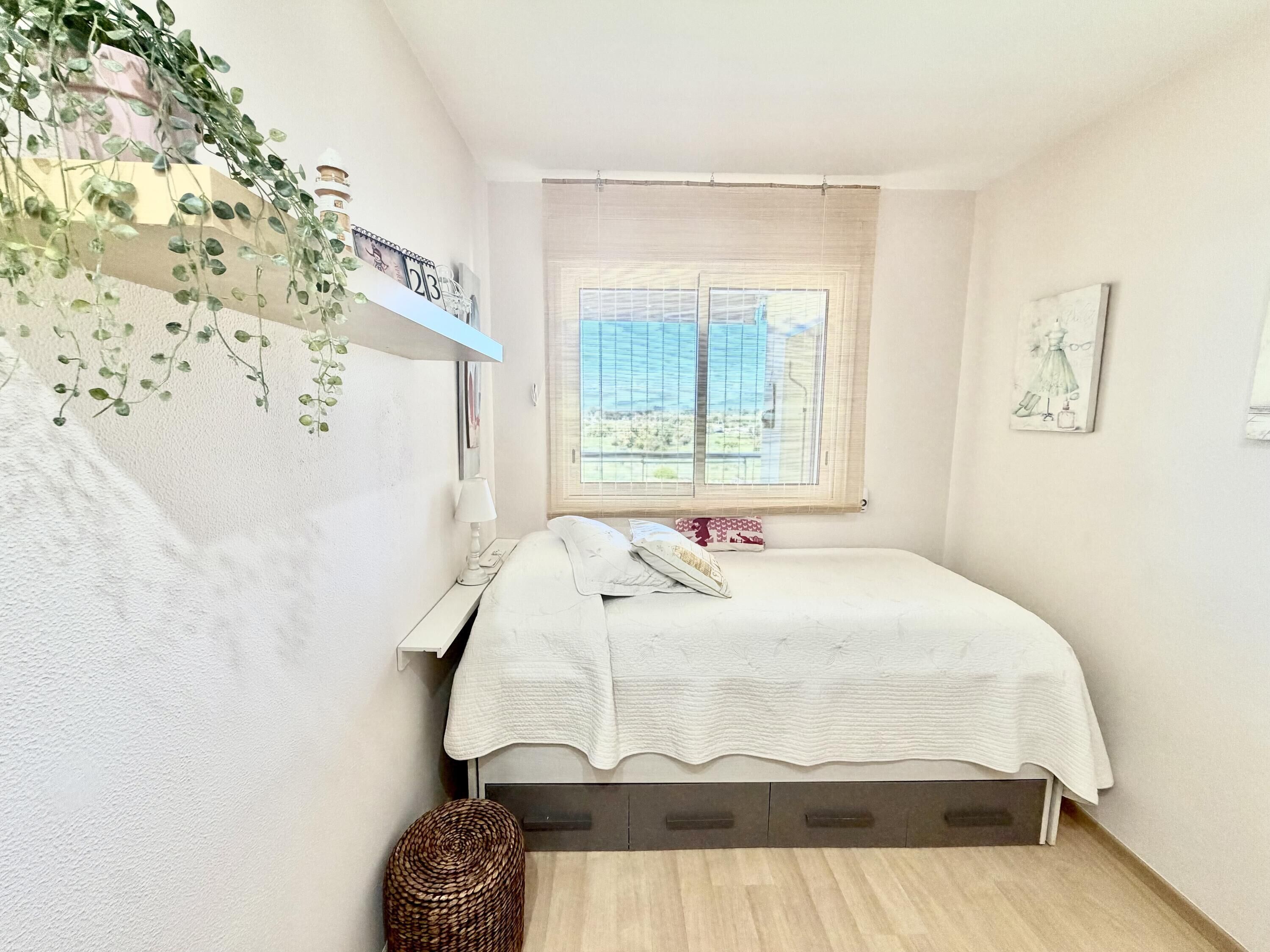 A bedroom with a bed, a window with blinds, a bedside table, a lamp, a wall-mounted shelf with a clock, and a woven basket.