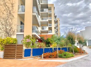 Un edificio residencial de varios pisos con balcones, entrada porticada y un jardín ajardinado.
