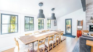 Un comedor con mesa y sillas de madera, lámparas colgantes y ventanas amplias.