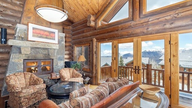 Interior de una cabaña de madera con chimenea de piedra, muebles con motivos decorativos y vistas a montañas a través de amplios ventanales.