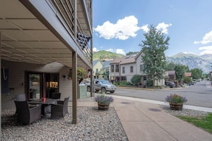 Ein Gebäude mit überdachter Terrasse, Sitzgelegenheiten im Freien und Straßenansicht mit Häusern und Bergen im Hintergrund.