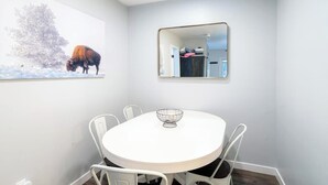 Un coin repas avec une table ronde, des chaises blanches, un panier en fil de fer, un tableau encadré représentant un bison et un miroir qui reflète une pièce avec une porte et un porte-manteau.