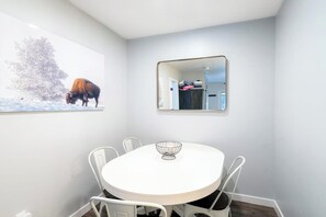 Ein Essbereich mit einem runden Tisch, weißen Stühlen, einem Drahtkorb, einem Bilderrahmen mit einem Bison und einem Spiegel, der einen Raum mit einer Tür und einem Garderobenhaken reflektiert.
