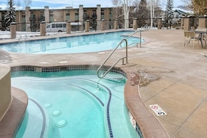 Ein Schwimmbad mit Whirlpool, eine Terrasse mit Stühlen und ein Gebäude im Hintergrund.