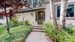 Una casa con puerta principal, escalones que la conducen y un jardín con diversas plantas y árboles.