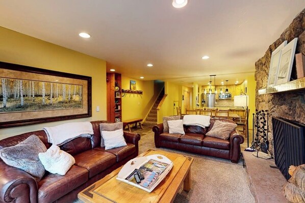 A living room with two leather sofas, a coffee table, a fireplace, and a staircase leading to a dining area. - Beaver Creek West Condo with Heated Pool, 3 Hot Tubs & Shuttle to the Slopes (Avon)
