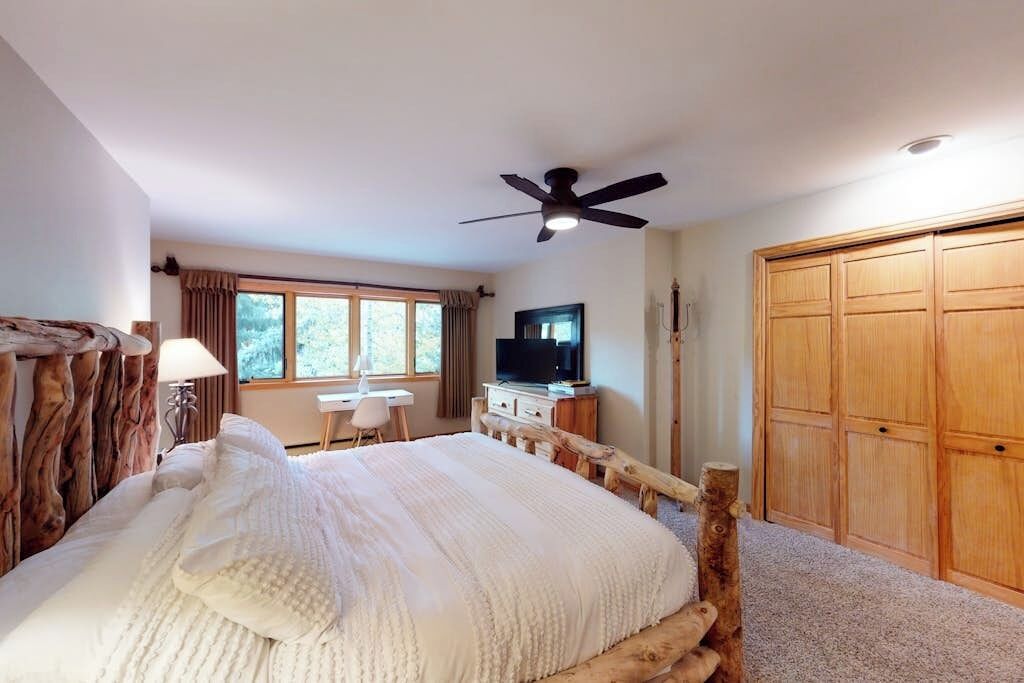 A bedroom with a wooden bed frame, a ceiling fan, a television, a desk, and a wardrobe.