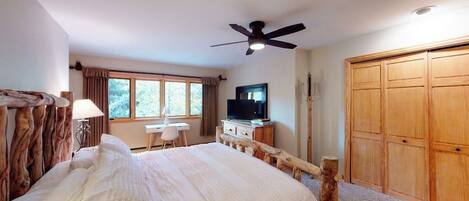 A bedroom with a wooden bed frame, a ceiling fan, a television, a desk, and a wardrobe.