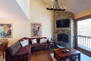 A living room with a stone fireplace, a television mounted above it, a dark-colored sofa, and a wooden coffee table. - Spacious Townhome with Hot Tub, Adjacent to Gore Creek | 4.5 Miles to Vail (Vail)