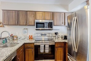 A kitchen with wooden cabinets, stainless steel appliances, a granite countertop, and a stainless steel refrigerator. - Golf Course Condo with Gas Fireplace, WiFi & Deck with Mountain Views (Avon)