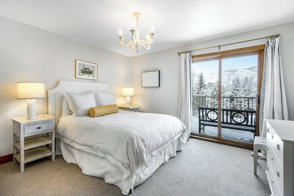 A bedroom with a bed, bedside tables, a chandelier, a window with a view of snow-covered trees, and a balcony.