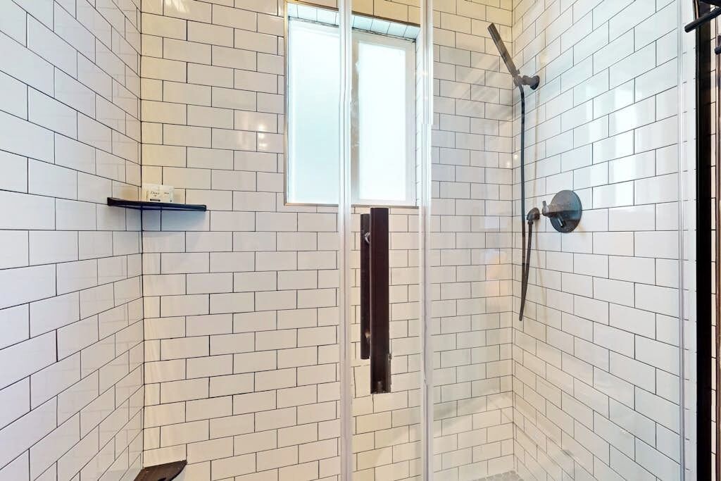 Une douche aux murs recouverts de carreaux blancs, une fenêtre et une étagère.