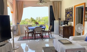A living room with a TV, a dining table with chairs, a sofa, a bookshelf, a ceiling fan, and a view of greenery outside. - Room-oasis Villa Private 1BR Luxury Pool (Karon)