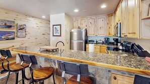 A kitchen with wooden cabinets, granite countertops, a stainless steel refrigerator, and bar stools. - Comfy condo w/hot tub, pool & sauna access- free shuttle to slopes, walk to lake (Avon)