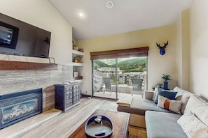 Ein Wohnzimmer mit Kamin, Flachbildfernseher, einer Couch und Blick auf einen Balkon mit Bergblick.
