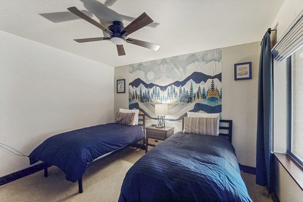 A bedroom with two beds, a ceiling fan, a mountain-themed wall mural, and a window with curtains.