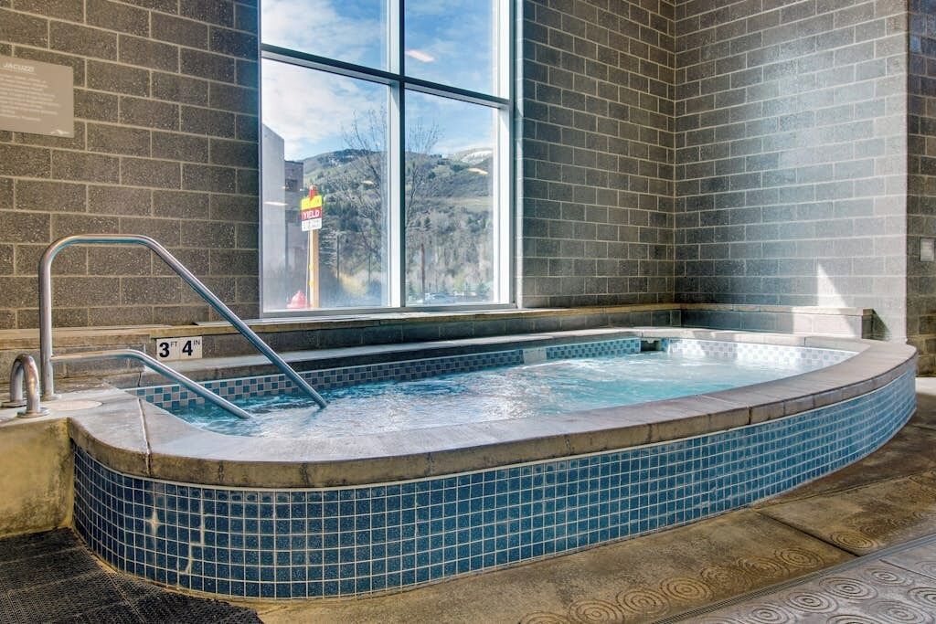 A hot tub with a tiled edge, a metal ladder, and a window with a view of a building and trees.