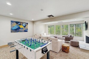 A modern living room with a foosball table, a beige sofa, a round ottoman, and a painting on the wall. - Inviting condo with large terrace, pool & hot tub - next to golf/Nordic skiing (Vail)