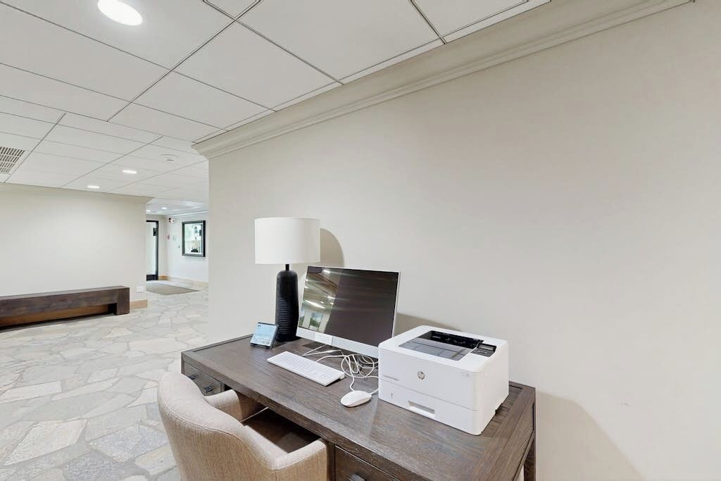 A hotel room with a desk, computer, printer, lamp, and a stone-patterned floor.
