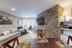Ein Wohnzimmer mit Steinwand, einem Flachbildfernseher, einer Couch, einem Esstisch mit Unterlagen und einem Gemälde an der Wand.