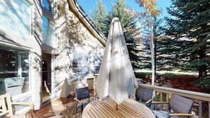 A deck with a round table and chairs, a covered umbrella, and a view of trees and a building. - Springs of Arrowhead Getaway w/ Deck, Pool, Hot Tub & Village Shuttle (Edwards)