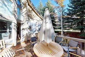 Eine Terrasse mit einem runden Tisch und Stühlen, einem Sonnenschirm und Blick auf Bäume und ein Gebäude.