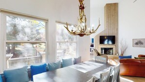 A dining area with a large table, chairs, and blue cushions. There is a fireplace with a TV above it, and a ceiling fan. - Springs of Arrowhead Getaway w/ Deck, Pool, Hot Tub & Village Shuttle (Edwards)