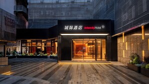Entrada de hotel moderna com fachada de vidro, sinalização em chinês e inglês, e área externa bem iluminada com plantas.