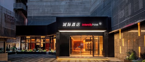 A modern hotel entrance with a glass facade, a sign in both Chinese and English, and a well-lit outdoor area with plants.