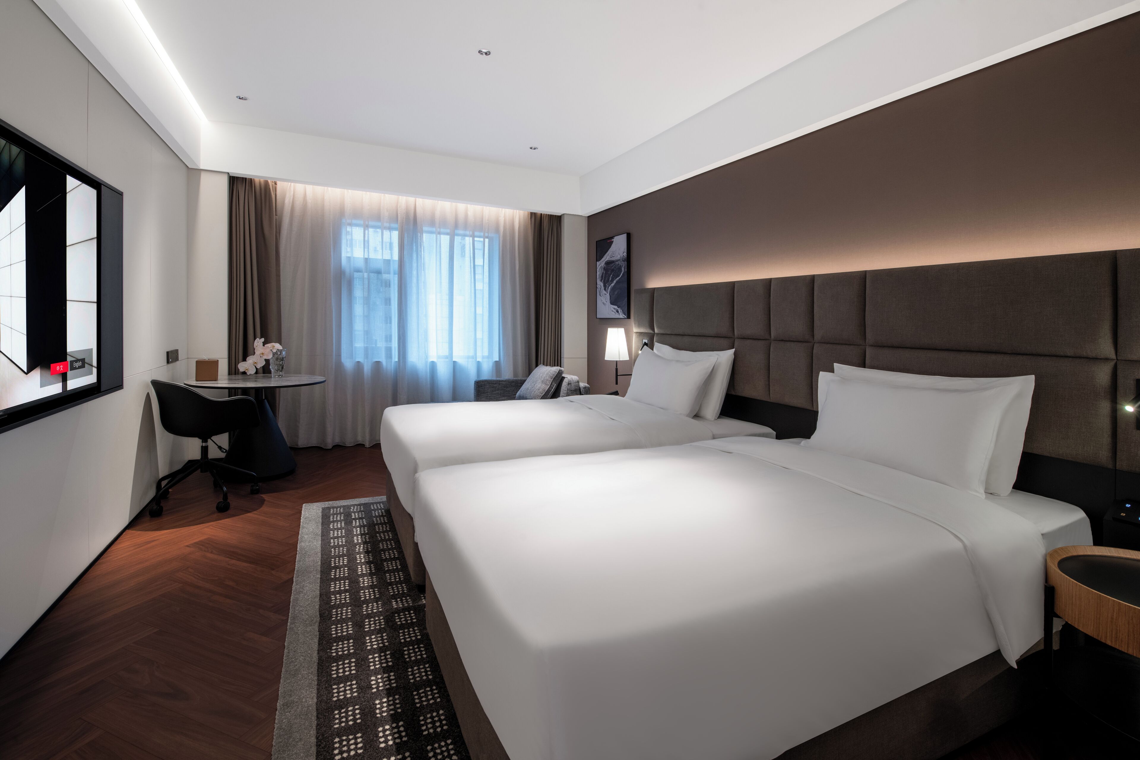 Ein Hotelzimmer mit zwei Betten, einem Schreibtisch, einem Stuhl, einer Lampe und einem großen Fenster mit Vorhängen.