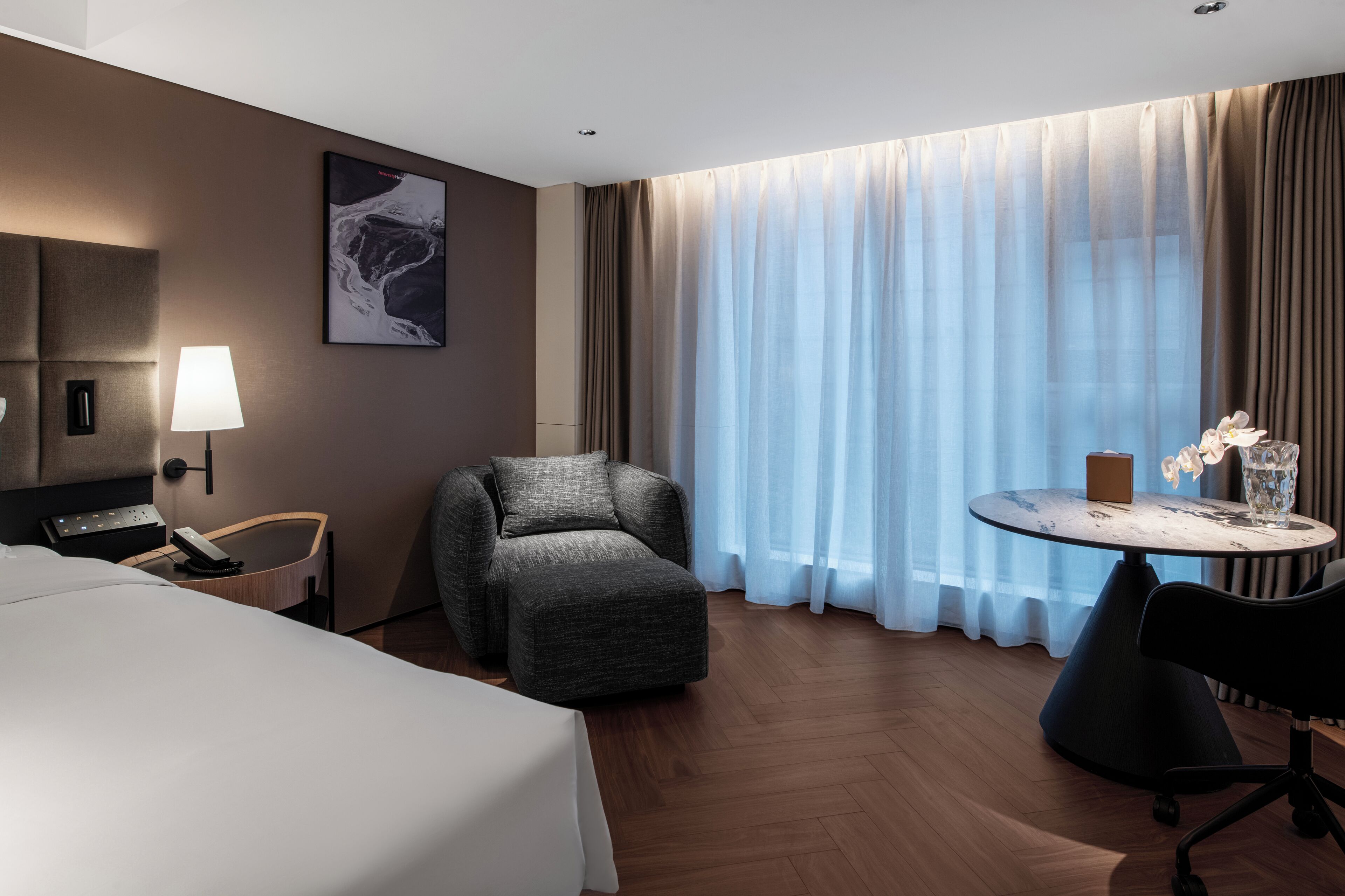 Une chambre d’hôtel avec un lit, un bureau, une chaise, une lampe, un tableau et une grande fenêtre avec des rideaux.