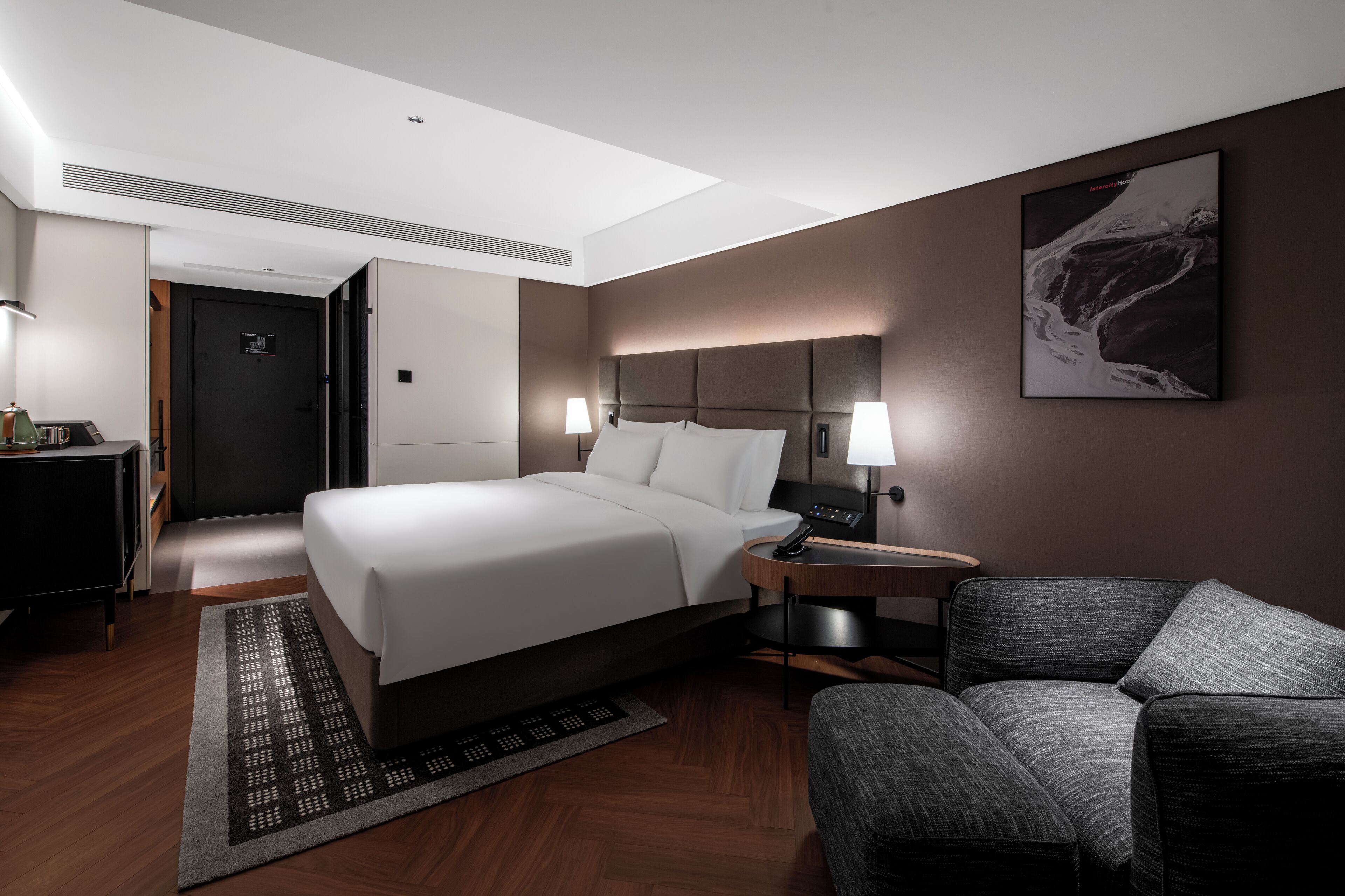 A modern hotel room with a bed, bedside table, lamp, and a chair.