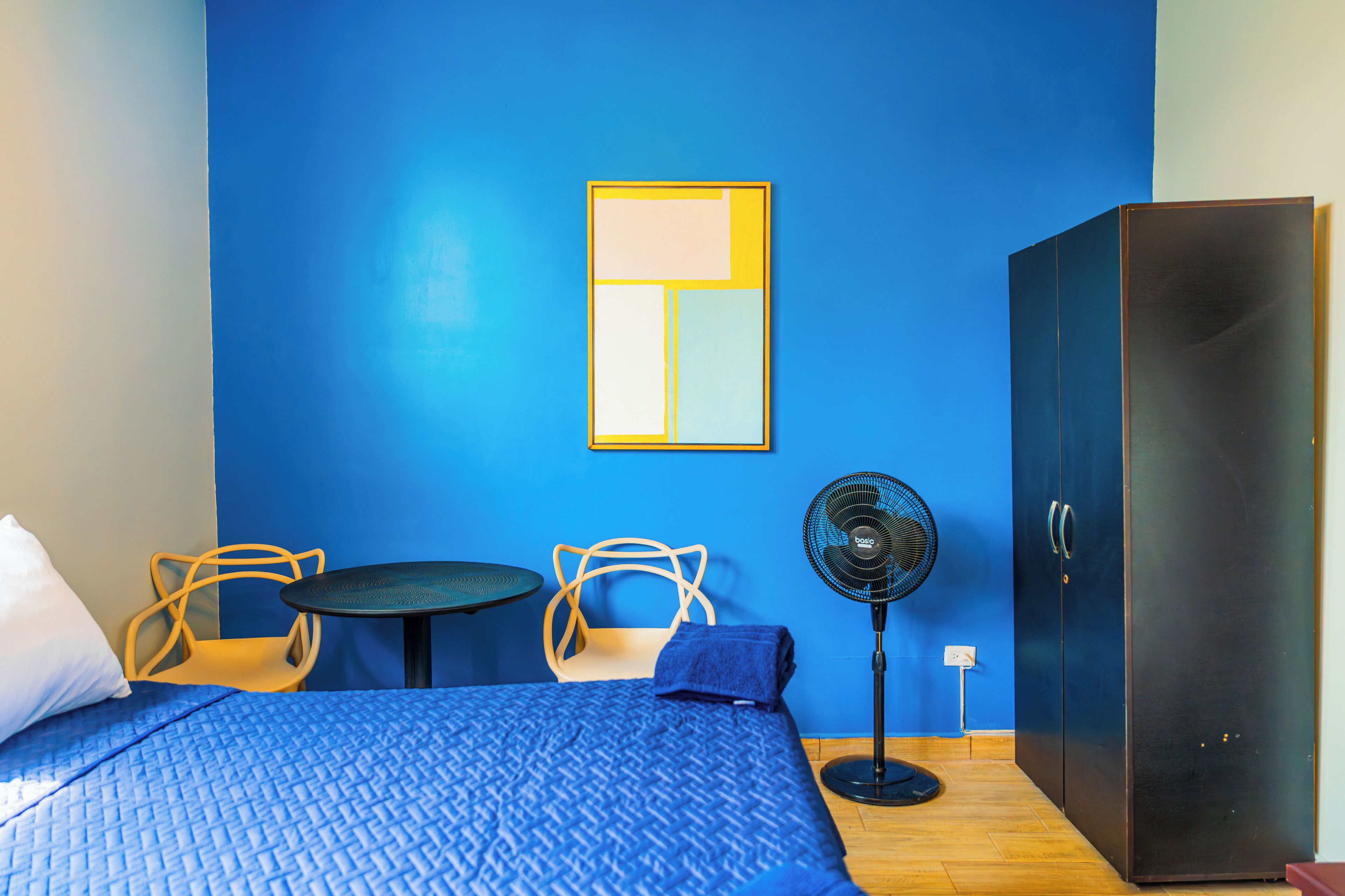Una habitación con una cama, dos sillas, una mesita, un ventilador y un armario.