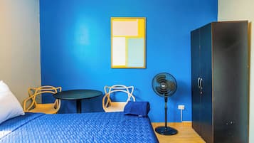 A room with a bed, two chairs, a small table, a standing fan, and a wardrobe.