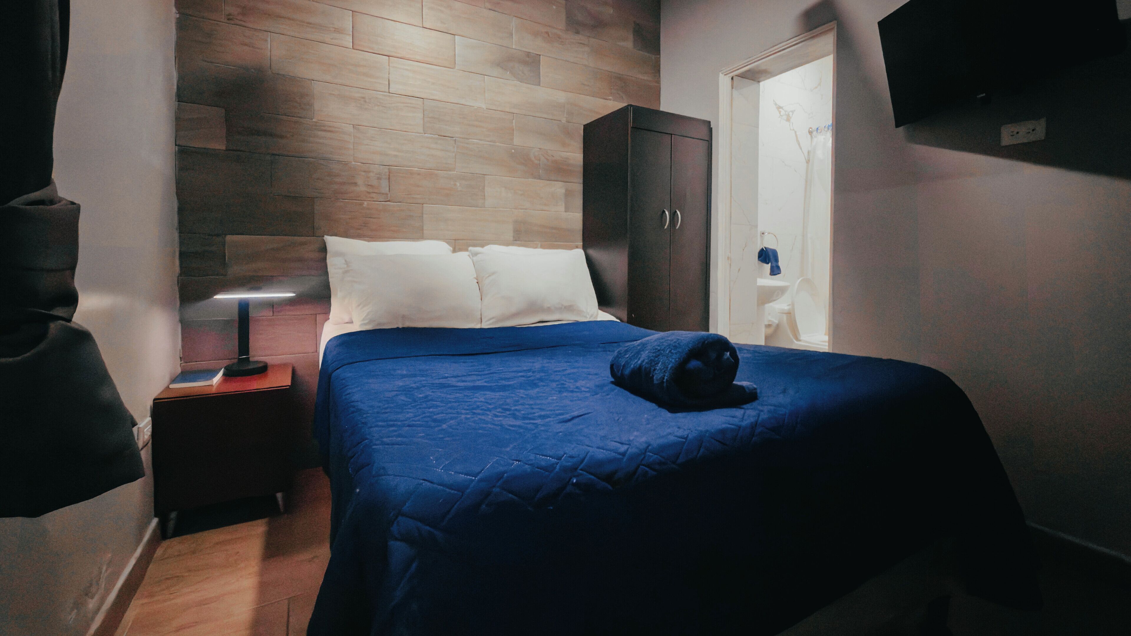 A bedroom with a wooden headboard, a bed with a blue bedspread, a nightstand with a lamp, and a wardrobe.