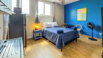 Un dormitorio con cama, mesita de noche, lámpara y ventilador de pie.